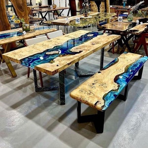 May include: A large wooden table with a blue and green epoxy resin river flowing through the center. The table has black metal legs and is paired with a matching bench. The table and bench are made of wood with a natural grain pattern.