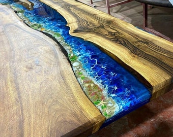 May include: A close-up of a wooden table top with a blue resin river running through it. The resin has white and blue swirls and includes small seashells and starfish.