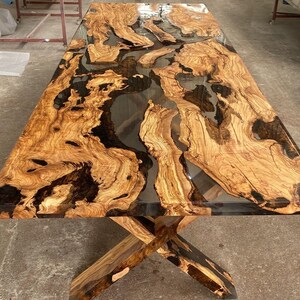 May include: A large wooden dining table with a clear epoxy resin top. The resin has a black and brown marbled pattern. The table has a simple X-shaped base.