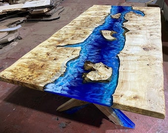 May include: A large wooden table with a blue epoxy river running down the center. The table has a natural wood finish and is supported by a simple X-shaped base.