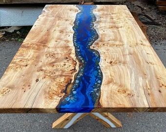 May include: A wooden table with a blue epoxy river running down the center. The epoxy contains white and blue seashells. The table has a natural wood finish and a white base.