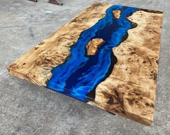 May include: A rectangular wooden table with a river of blue epoxy resin running through the center. The wood has a light brown color with dark brown knots and patterns. The epoxy resin is a vibrant blue.
