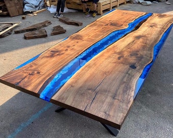 Puede incluir: Una mesa de madera grande con un río de resina epoxi azul que recorre el centro. La mesa tiene un acabado de madera natural y está sostenida por patas de metal negro.