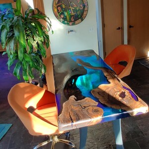 May include: A dark wood table with a blue and green epoxy resin top. The table has a unique, natural wood grain pattern and is supported by a single, curved metal leg. Two orange chairs are placed near the table.
