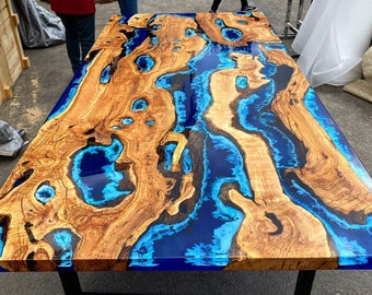 Op de afbeelding: Een houten tafelblad met blauwe en turquoise hars die de natuurlijke knoesten en scheuren in het hout vult. Het tafelblad is lichtbruin van kleur en de hars is diepblauw met een turquoise glans.