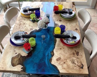 Pode incluir: Uma mesa de jantar retangular com um design de borda viva, apresentando um rio de epóxi azul. A mesa está posta com pratos brancos, copos coloridos e guardanapos pretos. A mesa tem aproximadamente 183 cm de comprimento.