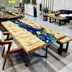 May include: A large wooden table with a blue and green epoxy resin river running through the center. The table has a black metal base and is surrounded by chairs.