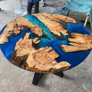 May include: A round wooden table with a blue epoxy resin top. The resin has a river-like design with a natural wood grain pattern. The table has a black metal base.