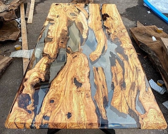 Op de afbeelding: Een groot houten tafelblad met een doorlopende rivier van heldere epoxyhars. Het hout is lichtbruin van kleur met donkerbruine knoesten en nerven. De hars is helder en heeft een lichte blauwe tint. Het tafelblad staat op een zwart metalen onderstel.