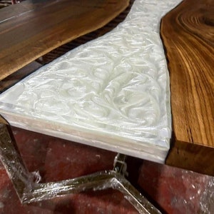 Puede incluir: Primer plano de una mesa de madera con un r&iacute;o de resina epoxi blanca que fluye por el centro. La resina tiene un patr&oacute;n de remolino y m&aacute;rmol. La mesa est&aacute; soportada por una base de metal.