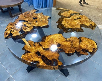 Könnte beinhalten: Runder Holztisch mit einer durchsichtigen Epoxidharzplatte. Das Holz hat eine natürliche braune und goldene Farbe mit einem einzigartigen Maserungsmuster. Der Tisch hat ein schwarzes Metallgestell mit einem kreuzförmigen Design.