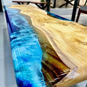May include: A wooden table top with a blue and white epoxy resin inlay that resembles a wave. The wood is a light brown color with darker brown streaks.