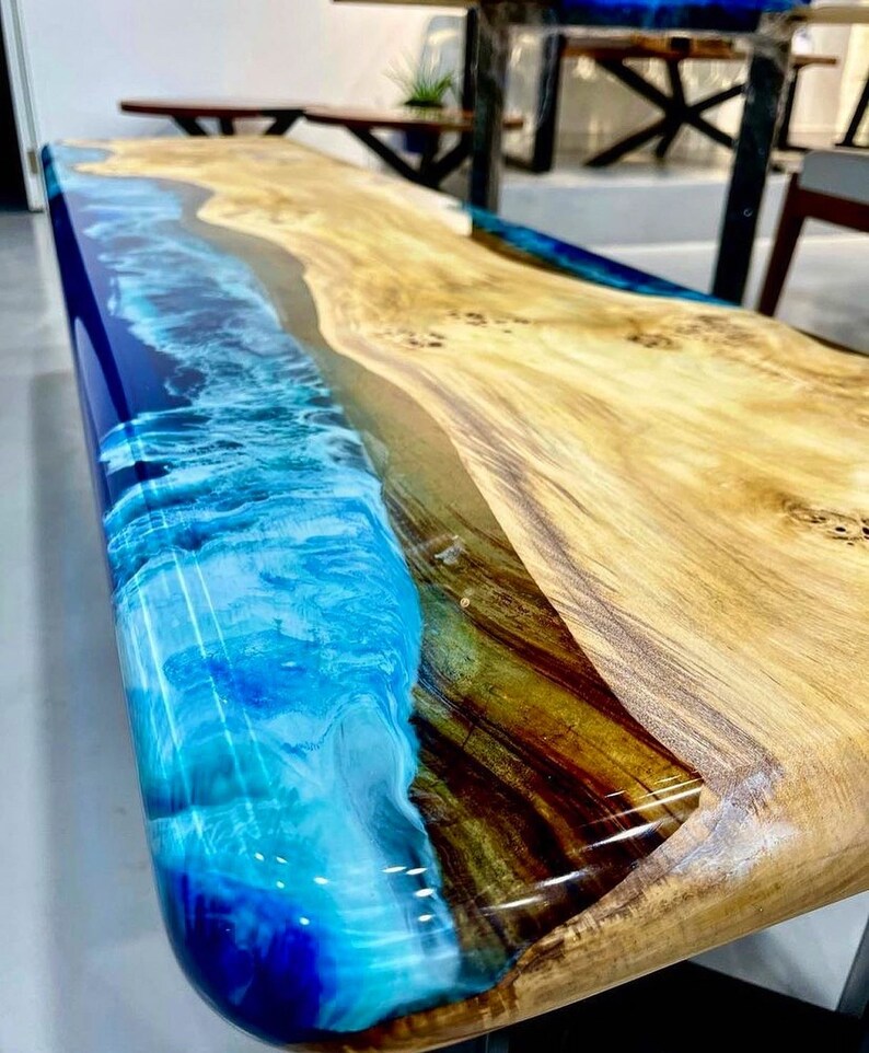May include: A wooden table top with a blue and white epoxy resin inlay that resembles a wave. The wood is a light brown color with darker brown streaks.