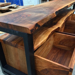 May include: A wooden console table with a live edge top and three drawers. The table is made of dark wood with a natural finish and has a black metal frame.