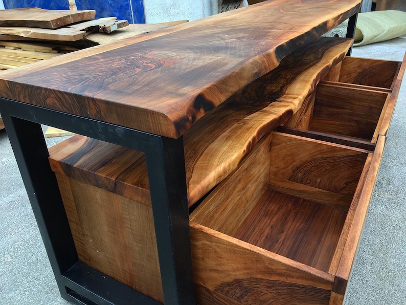 May include: A wooden console table with a live edge top and three drawers. The table is made of dark wood with a natural finish and has a black metal frame.