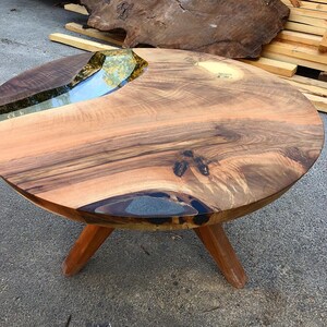 May include: Round wooden table with a natural wood top and a clear epoxy river running through the center. The table has three wooden legs.