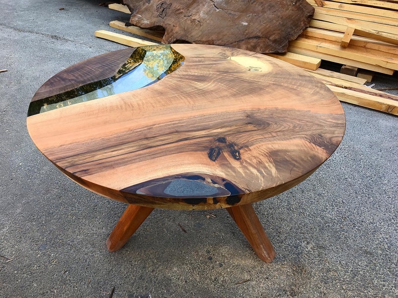 May include: Round wooden table with a natural wood top and a clear epoxy river running through the center. The table has three wooden legs.