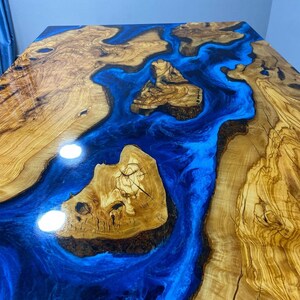 May include: A close-up of a wooden table top with a blue epoxy river running through it. The wood has a natural grain pattern and the epoxy is smooth and shiny.