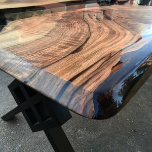 Puede incluir: Una mesa de madera con una tapa de borde vivo y un r&iacute;o de resina epoxi negra que atraviesa el centro. La mesa tiene una base de metal negro con un dise&ntilde;o geom&eacute;trico.