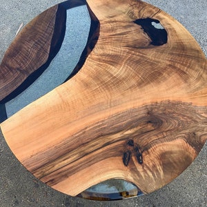 May include: Round wooden table top with a natural wood grain finish and a clear epoxy resin inlay. The inlay has a black border and a light blue center.