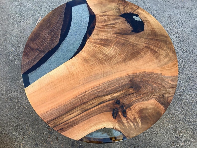 May include: Round wooden table top with a natural wood grain finish and a clear epoxy resin inlay. The inlay has a black border and a light blue center.