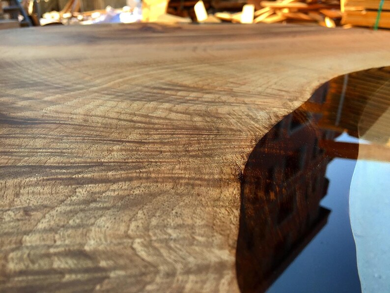 May include: Close-up of a wooden table top with a natural wood grain pattern. The edge of the table is filled with clear epoxy resin, creating a smooth, glassy surface.