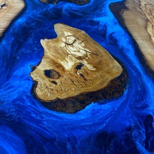 May include: A close-up of a wooden table top with a blue epoxy river running through it. The wood has a natural grain pattern and the epoxy has a swirling effect.
