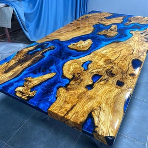 May include: A large wooden table top with a blue epoxy resin river running through it. The wood has a natural grain pattern and the resin is clear and glossy.