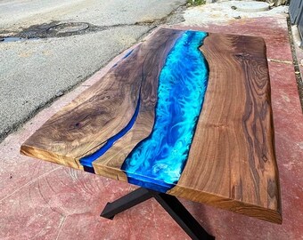 Puede incluir: Una mesa de madera con un río de resina epoxi azul que atraviesa el centro. La mesa está hecha de madera de nogal y tiene un acabado natural. El río de resina epoxi es transparente y tiene un ligero tono azul. La mesa está soportada por una base de metal negro.