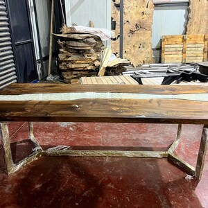 Puede incluir: Una gran mesa de comedor de madera con un r&iacute;o de epoxi blanco que recorre el centro. La mesa tiene una base de metal dorado.