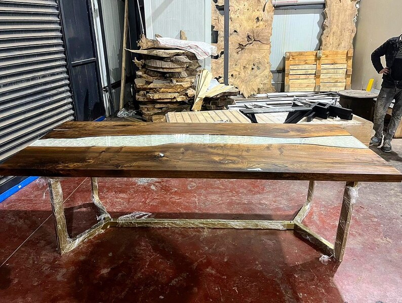 Puede incluir: Una gran mesa de comedor de madera con un r&iacute;o de epoxi blanco que recorre el centro. La mesa tiene una base de metal dorado.