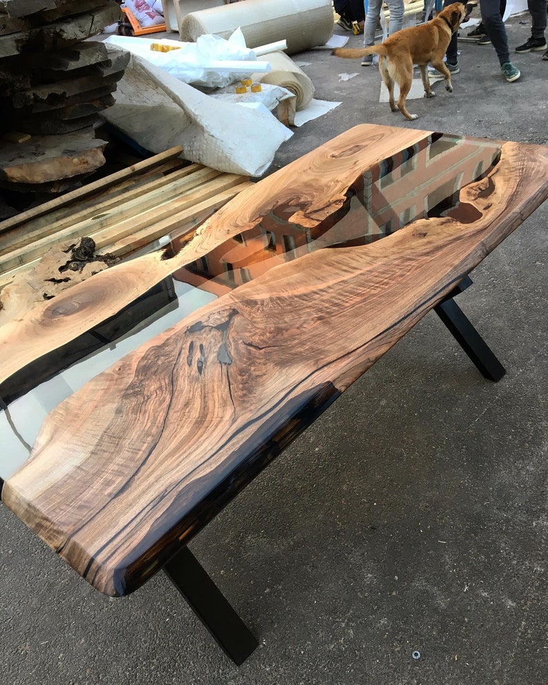 Puede incluir: Una mesa de madera con un borde vivo y un r&iacute;o de resina epoxi transparente que atraviesa el centro. La mesa tiene patas de metal negro.