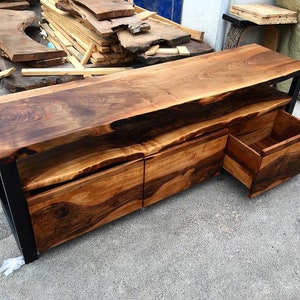 May include: A wooden entertainment center with a live edge top and three drawers. The drawers are made of wood with a dark brown finish. The entertainment center is made of metal with a black finish.