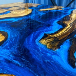 May include: Close-up of a blue and brown epoxy resin river table. The resin has a swirling, flowing pattern, resembling water. The wood is dark brown with a natural grain.