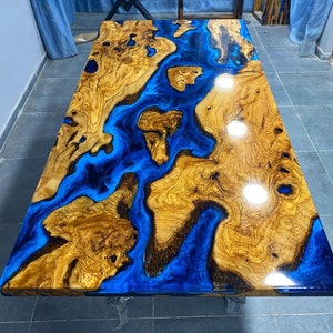 May include: A wooden table top with a blue epoxy resin inlay. The resin has a flowing, river-like design. The wood is a rich brown color with a natural grain pattern.