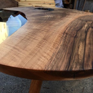 May include: A round wooden table with a live edge and a clear epoxy river running through the center. The table has a natural wood finish and is supported by a single wooden leg.