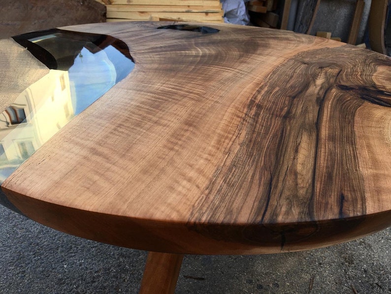 May include: A round wooden table with a live edge and a clear epoxy river running through the center. The table has a natural wood finish and is supported by a single wooden leg.