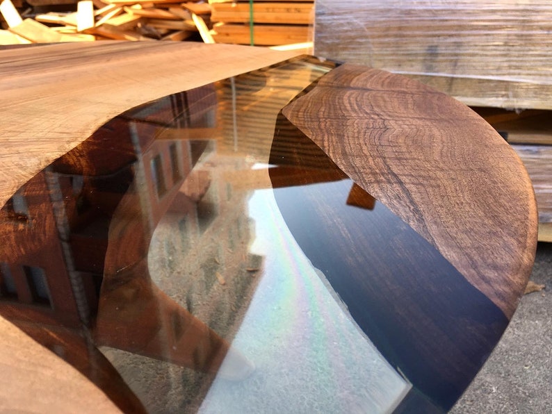 May include: Close-up of a wooden table top with a clear epoxy resin inlay. The resin is a dark blue color and has a wavy shape. The wood is a rich brown color and has a natural grain pattern.