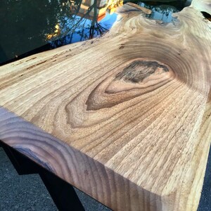 Puede incluir: Primer plano de una mesa de madera con un r&iacute;o de resina epoxi negra que la atraviesa. La madera tiene un veteado natural y la resina es lisa y brillante.