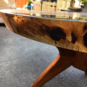 May include: Close-up of a round wooden table with a clear epoxy resin top. The table has a natural wood finish with dark brown accents and a unique, irregular edge. The table is supported by three wooden legs.