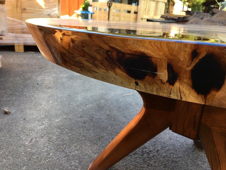 May include: Close-up of a round wooden table with a clear epoxy resin top. The table has a natural wood finish with dark brown accents and a unique, irregular edge. The table is supported by three wooden legs.