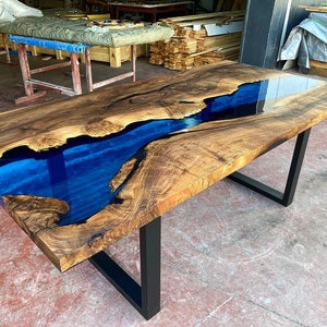 Op de afbeelding: Een grote houten eettafel met een blauwe epoxyrivier die door het midden loopt. De tafel heeft een zwart metalen onderstel.