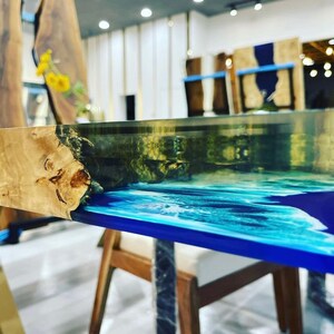 May include: Close-up of a wooden table top with a blue and white epoxy resin inlay. The table top is made of natural wood with a unique grain pattern. The epoxy resin is clear and has a smooth, glossy finish.