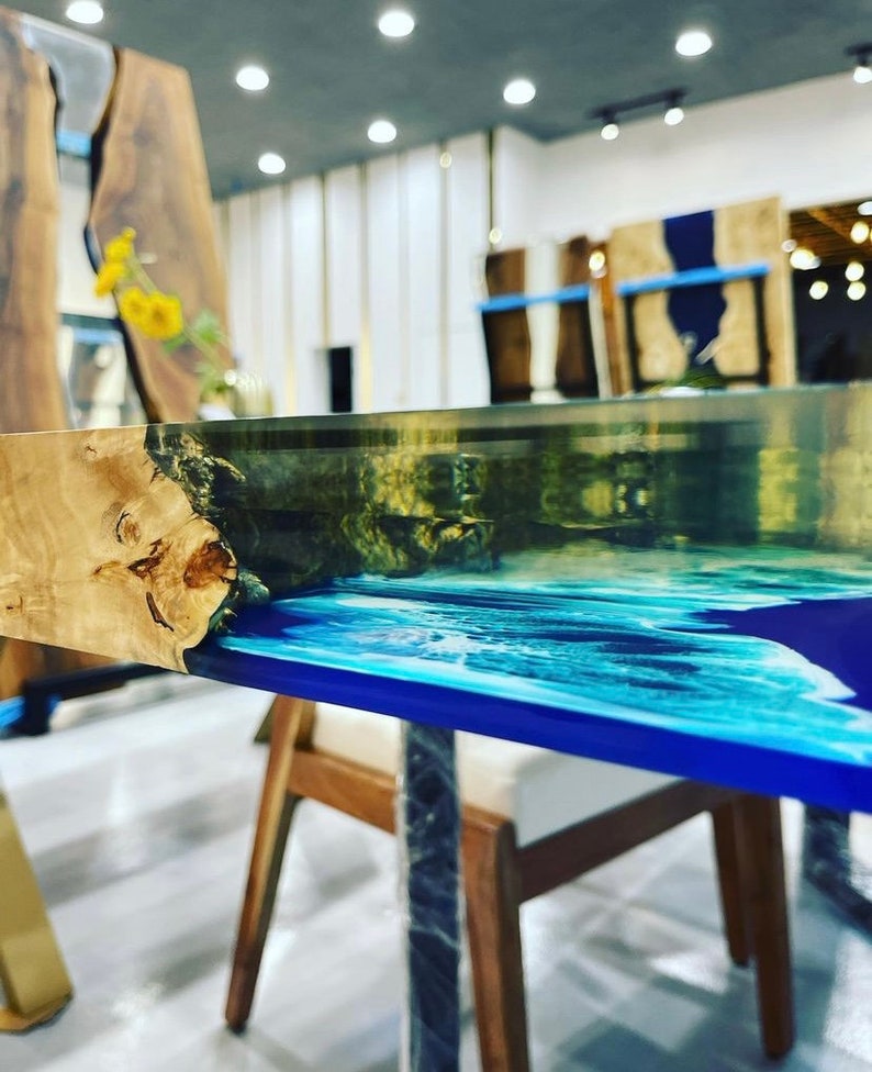 May include: Close-up of a wooden table top with a blue and white epoxy resin inlay. The table top is made of natural wood with a unique grain pattern. The epoxy resin is clear and has a smooth, glossy finish.