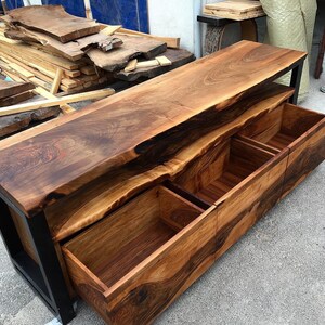 May include: A wooden entertainment center with a live edge top and three drawers. The drawers are made of wood and have a natural finish. The entertainment center is supported by a black metal frame.