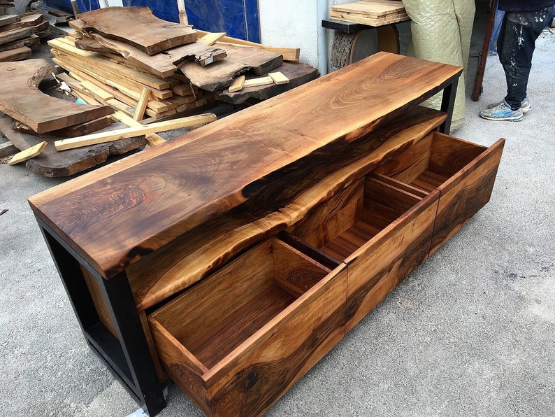 May include: A wooden entertainment center with a live edge top and three drawers. The drawers are made of wood and have a natural finish. The entertainment center is supported by a black metal frame.