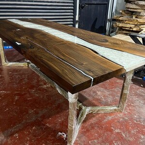 Puede incluir: Una mesa de comedor grande de madera oscura con un r&iacute;o de resina blanca que recorre el centro. La mesa tiene un dise&ntilde;o moderno e industrial con patas de metal dorado.