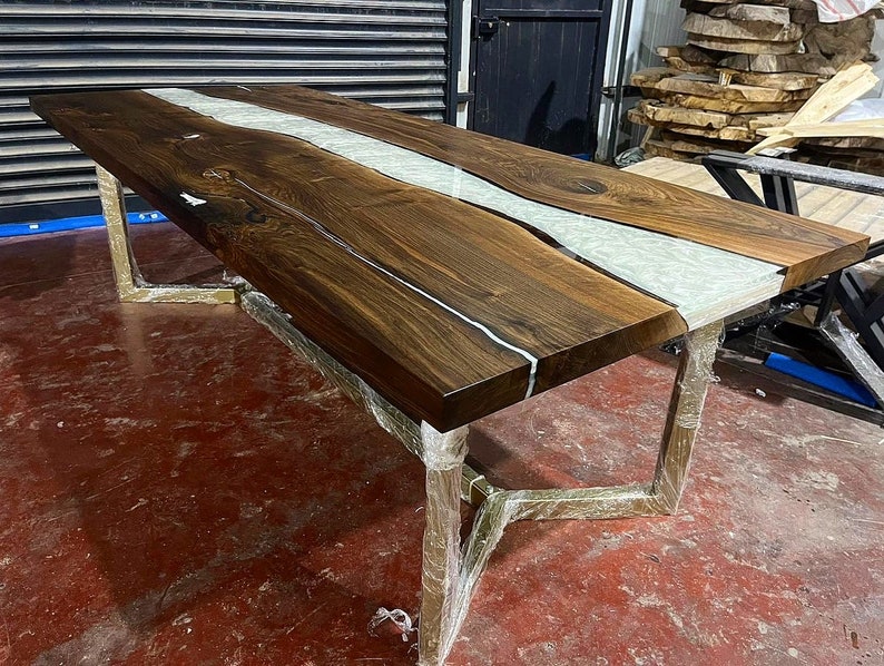 Puede incluir: Una mesa de comedor grande de madera oscura con un r&iacute;o de resina blanca que recorre el centro. La mesa tiene un dise&ntilde;o moderno e industrial con patas de metal dorado.