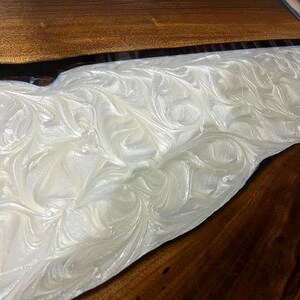 Puede incluir: Primer plano de una mesa de madera con un r&iacute;o de resina epoxi blanca que fluye por el centro. La resina tiene un patr&oacute;n de remolino y m&aacute;rmol.