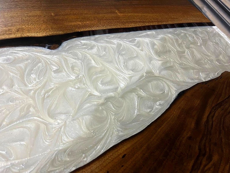 Puede incluir: Primer plano de una mesa de madera con un r&iacute;o de resina epoxi blanca que fluye por el centro. La resina tiene un patr&oacute;n de remolino y m&aacute;rmol.